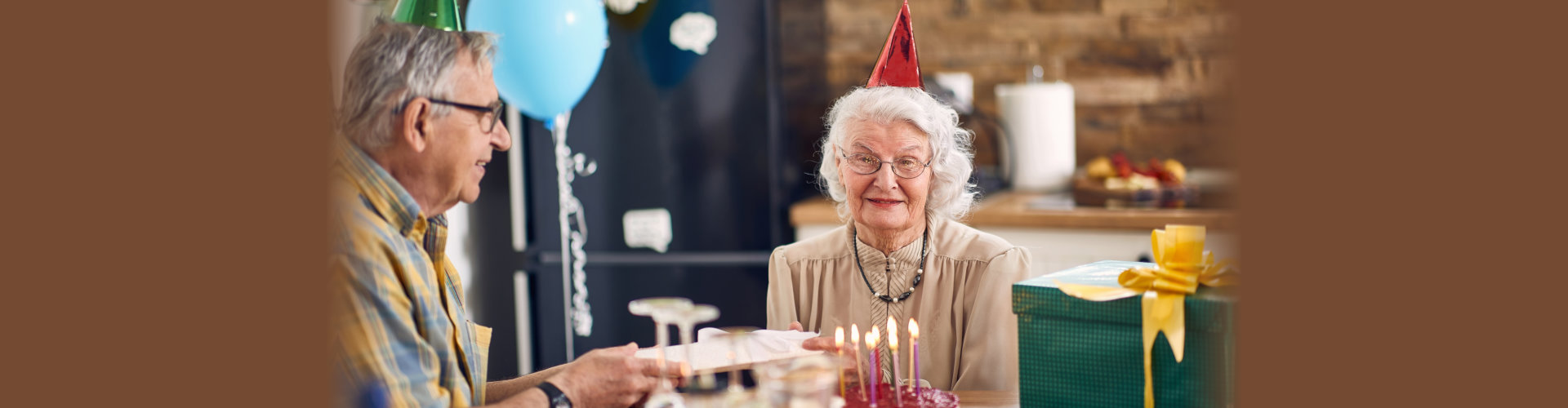 Beautiful happy senior couple sitting at kitchen table in birthday hats, elderly man giving a birthday present to her wife. Lifestyle, senior life, togetherness concept.